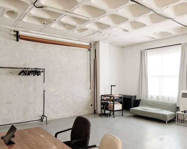 The image shows a minimalist studio space with gray walls, a large window, a light-colored sofa, a clothing rack, and an air conditioning unit.