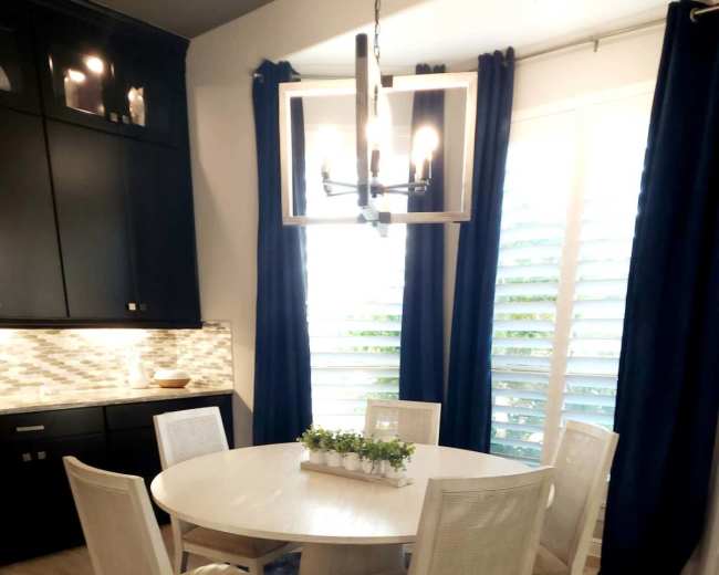 A round dining table with six chairs is set up in a well-lit area with dark cabinetry and blue curtains.