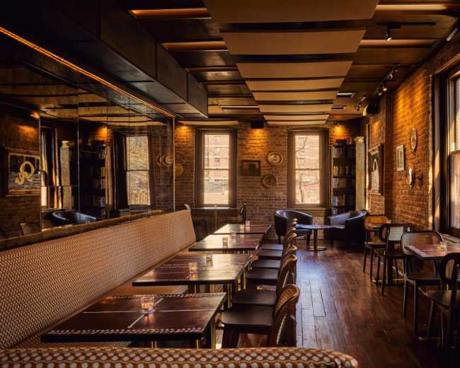 The image shows a warm, inviting restaurant interior with brick walls, wooden floors, and a mix of seating options including tables and chairs along with a long banquette.