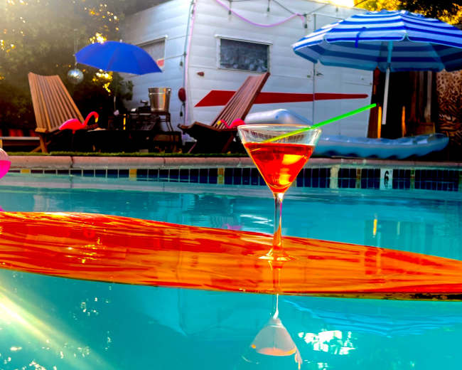 A cocktail glass rests on an orange surfboard in a swimming pool surrounded by colorful umbrellas and lounge chairs.