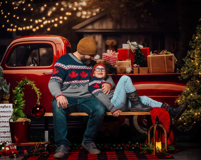 A man and a girl sit together on a bench in front of a vintage red truck decorated for Christmas, surrounded by presents and a brightly lit tree.