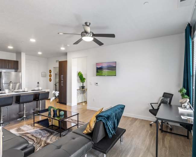 The image shows a modern living room featuring a black sofa, a coffee table, a flat-screen TV mounted on the wall, and a desk in the corner, with a kitchen visible in the background.