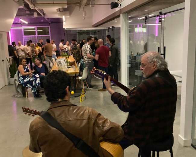 A small crowd gathers to listen to two musicians playing guitars in a contemporary indoor space.