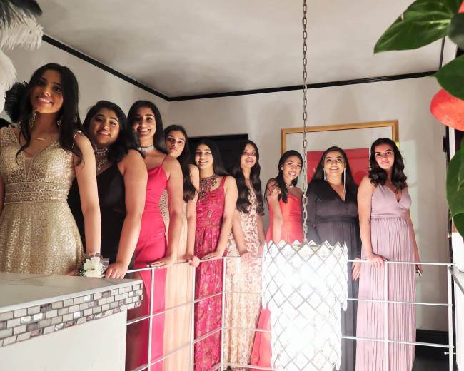 A group of ten women in formal dresses poses together near a decorative light fixture in a stylish indoor setting.