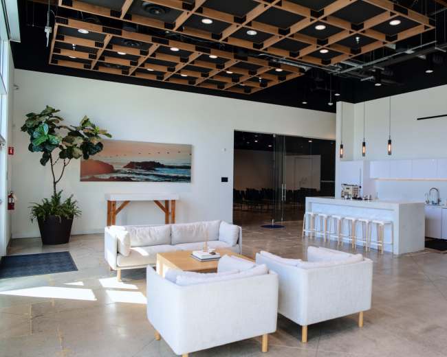 The image shows a modern lobby area featuring a seating arrangement with light-colored couches, a kitchen space with barstools, and large windows allowing natural light to enter.