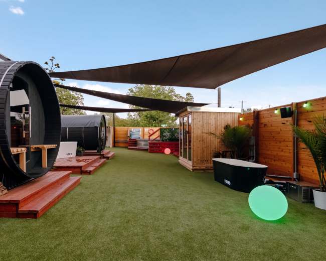 The image shows a landscaped outdoor area featuring two barrel-shaped structures, wooden platforms, and green turf, all shaded by large fabric canopies.