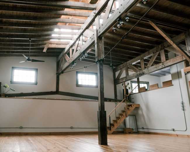 The image shows an interior space with high ceilings, exposed wooden beams, and a staircase leading to an upper level.