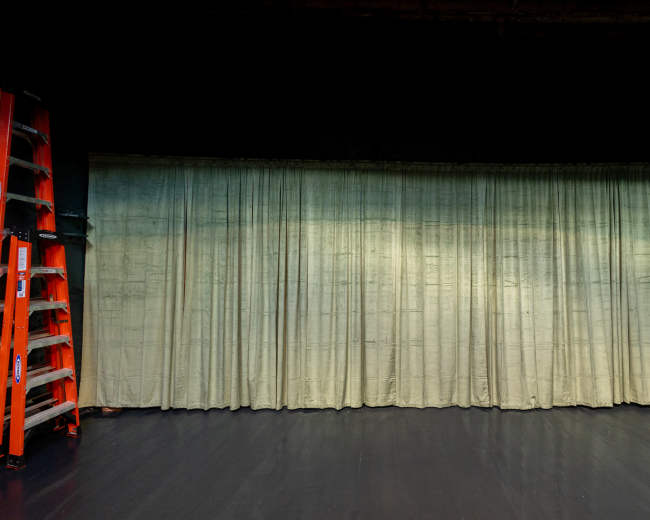 A green curtain hangs in a dimly lit room next to an orange ladder.