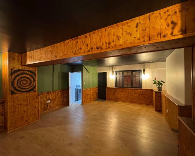 The image shows a spacious interior room featuring wood paneling, a dark ceiling, and a doorway leading to another area, with a plant and a patterned design on one wall.