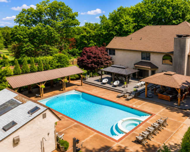 The image shows a spacious backyard featuring a large swimming pool, surrounded by lounge chairs and shaded areas, with a house and trees in the background.