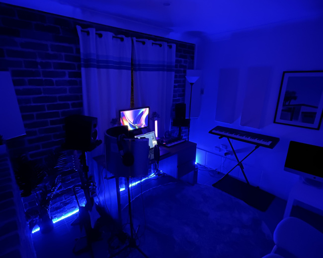 A dimly lit room with blue lighting, featuring music equipment, a computer, and a keyboard against a brick wall.