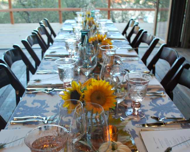 A long dining table is set with glasses, cutlery, and decorations including sunflowers and a small pumpkin.