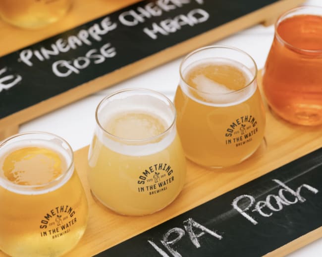 A wooden tasting board displays several glasses of craft beer, each labeled with the names of the different varieties including "Light," "IPA," and "Peach."