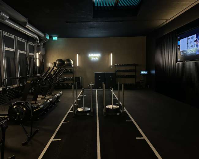 The image depicts a modern gym studio featuring workout equipment, including treadmills and weights, with a minimalist design and dim lighting.
