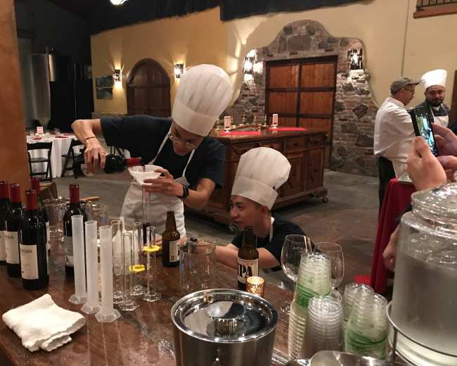 Two chefs wearing white hats are preparing drinks at a bar surrounded by bottles of wine and beer in a rustic setting.