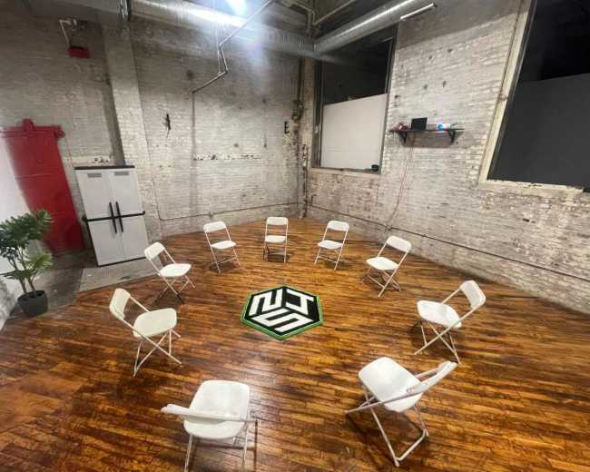 A circular arrangement of white chairs is set on a wooden floor in a sparse, industrial-style room with exposed brick walls and a visible fridge.