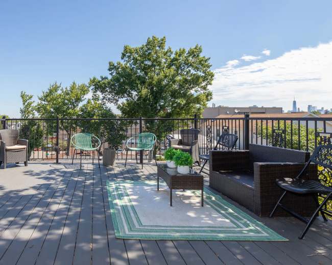 The image shows a rooftop terrace with various seating options and a striped rug, surrounded by trees and buildings in the background.