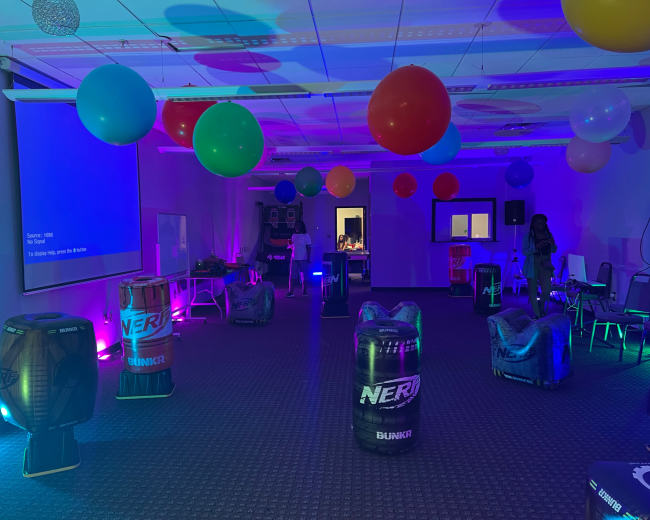The room is set up for an event, featuring colorful balloons and a series of Nerf-themed obstacles arranged around a carpeted floor.