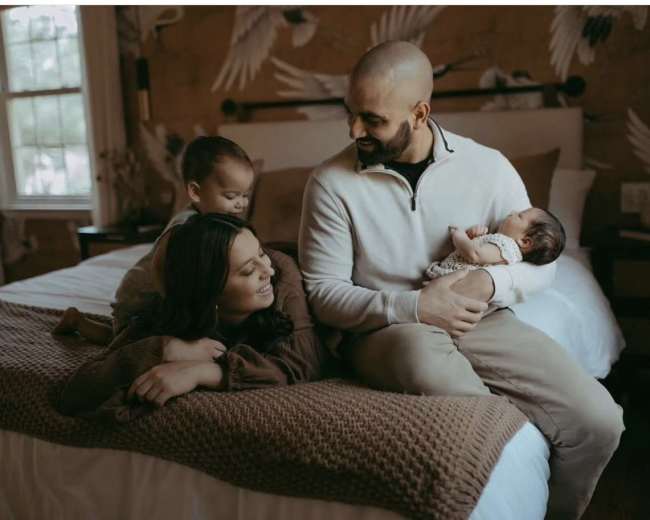 A family sits together on a bed, with two children interacting and a parent holding an infant.
