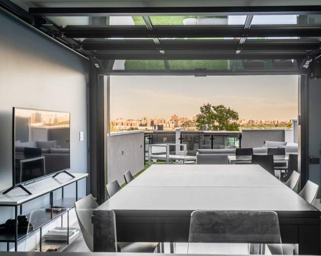 A modern dining area with a large table and a retractable glass wall revealing a city skyline view.