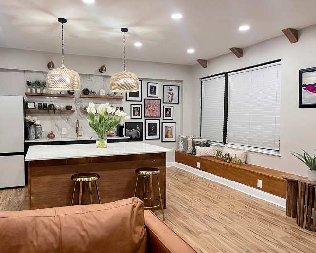 A modern kitchen features a white marble island with bar stools, framed artwork on the walls, and a vase of tulips, with natural light coming through the windows.
