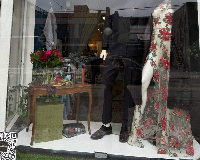 A clothing store display features two mannequins, one dressed in a formal black outfit and the other in a floral-patterned dress, alongside decorative items and flowers.