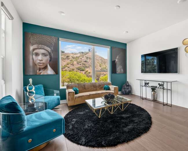 The living room features a modern design with a gold sofa, turquoise chairs, a black area rug, and large windows displaying a hillside view.