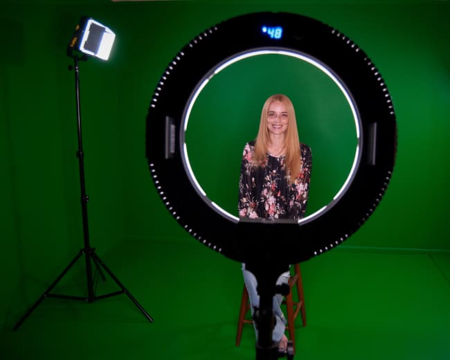 A woman sits on a stool in front of a green screen, framed by a circular light, with studio lights positioned on either side.