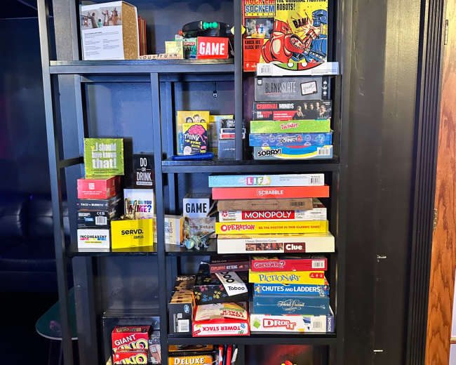 The image shows a black bookshelf filled with various board games and colorful game boxes, displayed alongside a few decorative items.