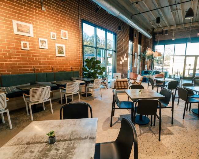 The image shows a modern café interior with brick walls, wooden ceilings, and a mix of tables and chairs arranged around the space.