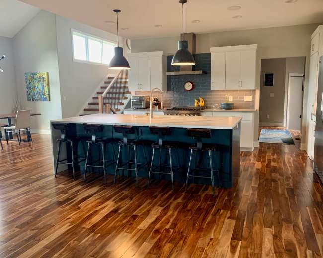 The image shows a modern kitchen with a central island, bar stools, and stainless steel appliances, featuring hardwood floors and large windows.