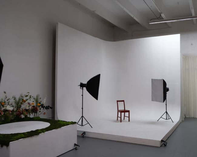 The image shows a minimalist photography studio with a single wooden chair positioned on a white platform, flanked by two softbox lights and a floral arrangement on one side.
