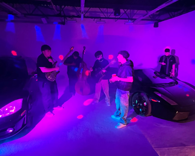 A group of musicians is playing instruments near two luxury cars in a dimly lit space with purple and blue lighting.