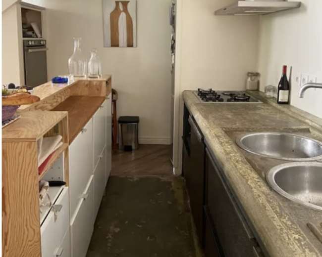 The image shows a narrow kitchen area with a counter on the left, a stove and sink on the right, and stairs leading up in the background.