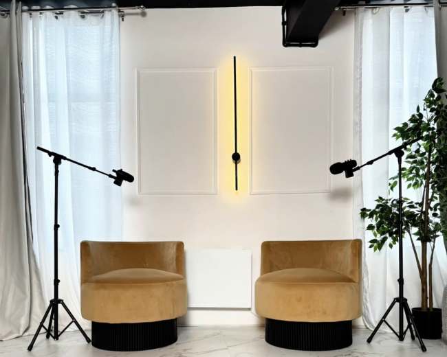 The image shows a modern interior space featuring two yellow chairs positioned across from a white wall with a vertical light fixture, flanked by two standing microphones and a potted plant.