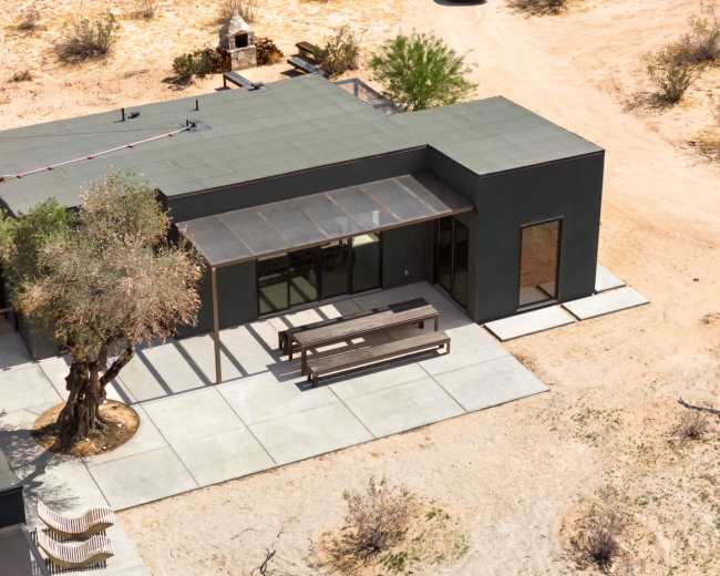 A modern, single-story house with a flat roof and large windows sits on a dry, sandy landscape, featuring a shaded outdoor seating area and a nearby olive tree.