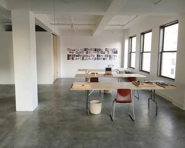 An empty, minimalist workspace features wooden tables and chairs, with a wall adorned with a collection of photographs.