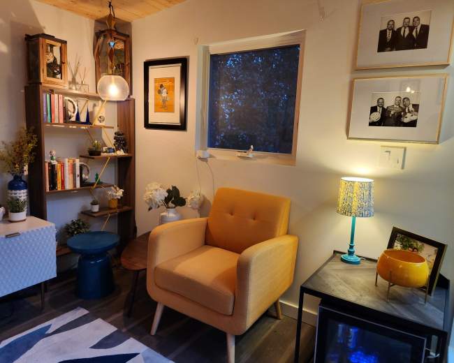 A cozy living room corner with a yellow armchair, bookshelf filled with books and decor, a side table, and a lamp.