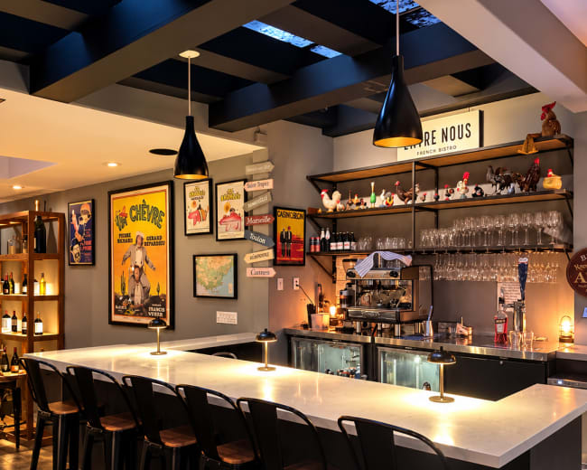 The image shows a modern bar area with a sleek white countertop, black metal stools, and shelves displaying various bottles and decor.