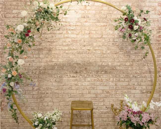 A circular floral arrangement on a brick wall, with a gold stand and a stool positioned in front of it.