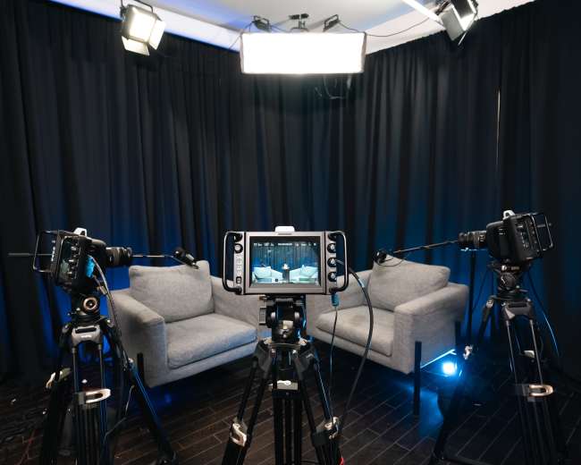 Two gray sofas are positioned in front of multiple cameras and lights, set against a black backdrop in a studio environment.
