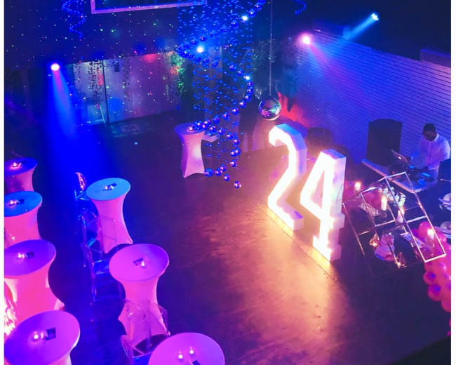 A decorated event space features illuminated tables and a large "24" display under colorful lights, with a DJ setup in the corner.
