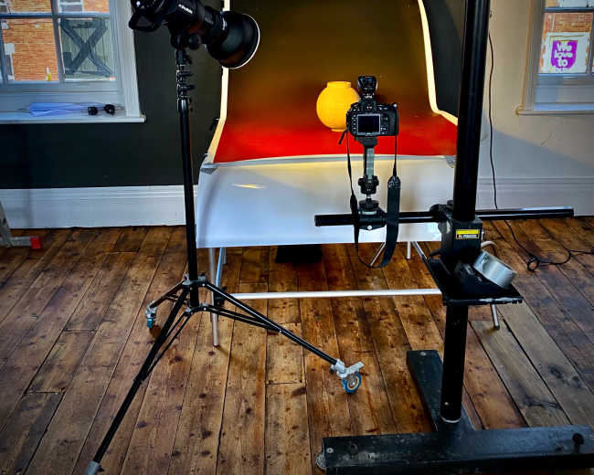 A photography setup features a camera and light positioned in front of a curved white backdrop, illuminating a round object on a red surface in a room with wooden floors.