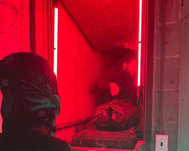 A person wearing a hooded garment stands in front of a brightly lit opening in a concrete wall, while another figure is seen inside the opening.