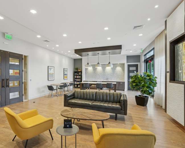 The image shows a modern lobby area with a black leather sofa, two yellow chairs, a coffee table, and a dining area in the background.