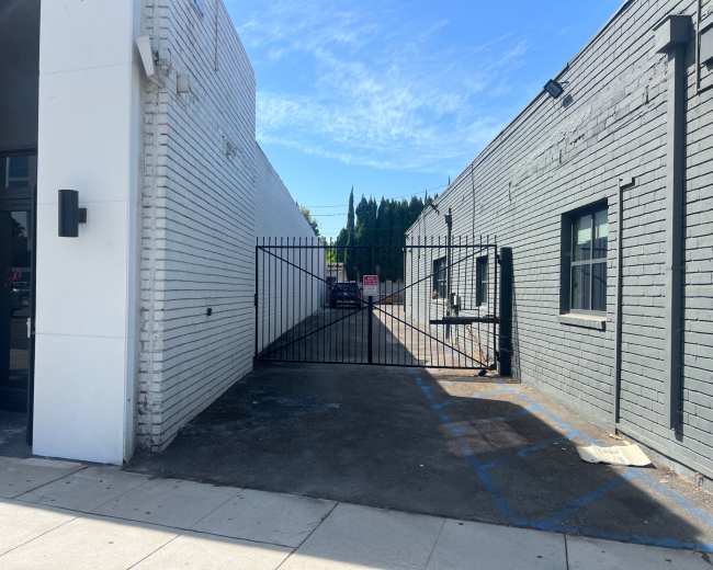 A narrow alleyway with a gated entrance is flanked by two buildings, one white and the other gray, under a clear blue sky.