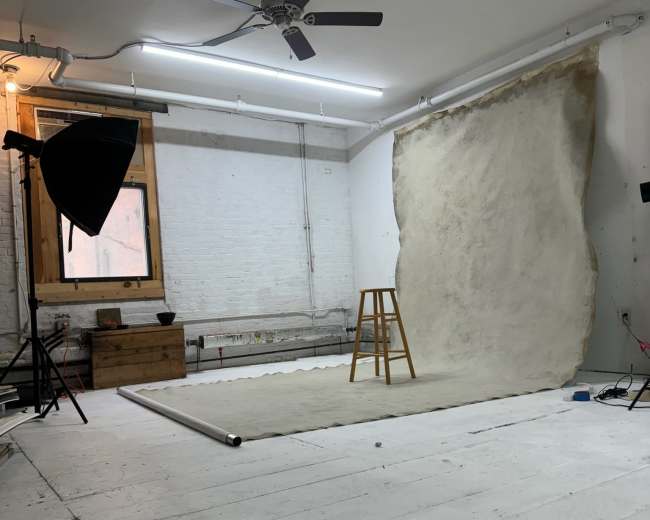 A photography studio is set up with a wooden stool, a backdrop partially rolled out, and lighting equipment arranged around a whitewashed room.