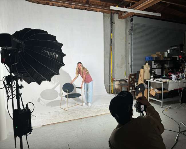 A photographer captures a model adjusting a chair in a studio with a backdrop and various equipment visible in the background.