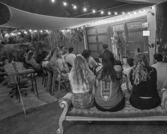 A group of people sits on a plush couch, watching a performer on stage in a backyard illuminated by string lights.