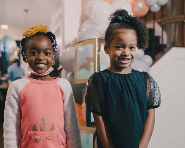 Two young girls stand smiling in a decorated indoor space, with balloons in the background.